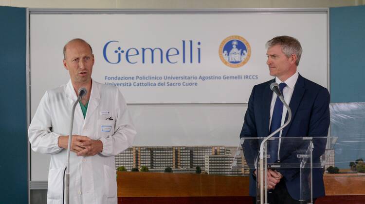 dr sergio alfieri wearing a white labcoat speaks next to matteo bruni who wears a navy suit. they stand in front of a sign that reads "gemelli hospital"