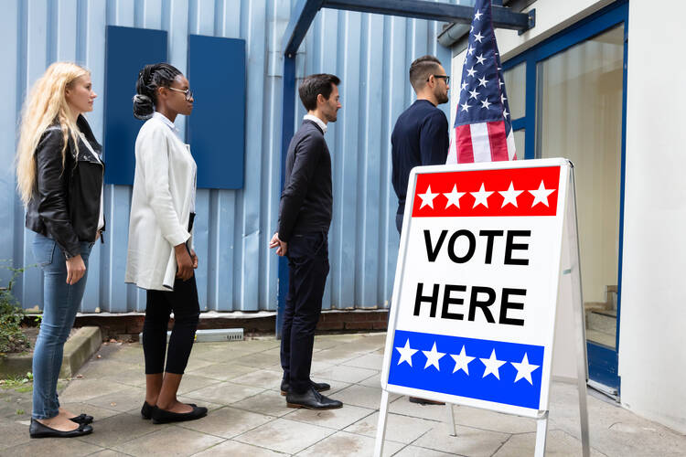 Young men and women stand in line outside a polling place with a "Vote Here" sign.